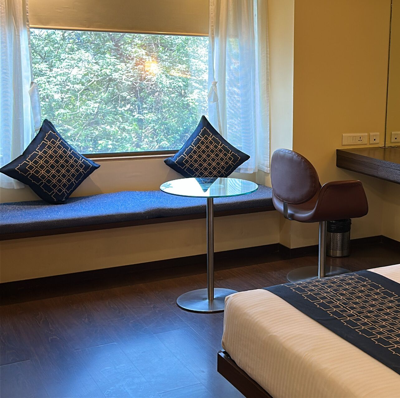 A room with a bed, a round glass table, two blue pillows, a brown chair, and a window with a view of trees.