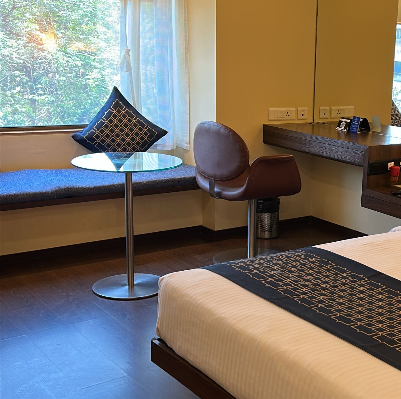 A hotel room with a bed, a round glass table with a single chair, a window with a view of trees, and a small desk with a phone and a red cup.