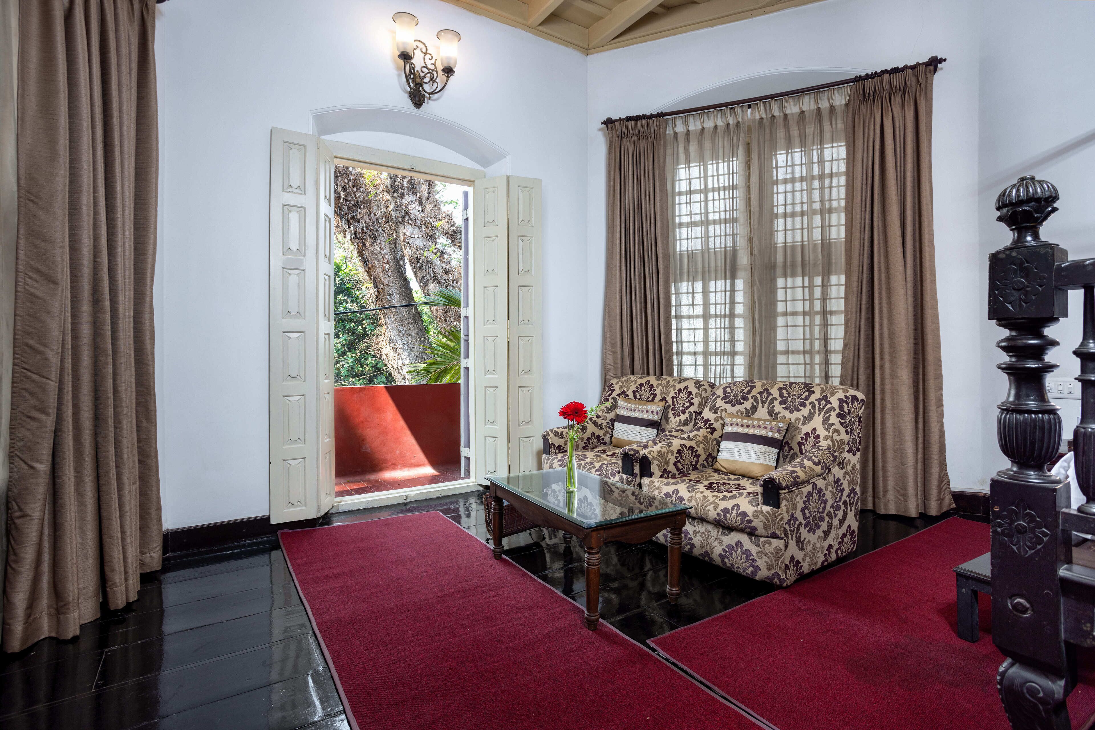 A room with a patterned sofa, a glass coffee table with a vase, a red rug, and a view of greenery through a door.