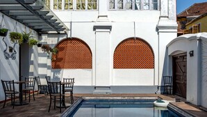 A pool area with a clear view of the pool, a patio with chairs, and a building with large windows and arched openings. - Koder House Heritage Hotel & Spa (Kochi)