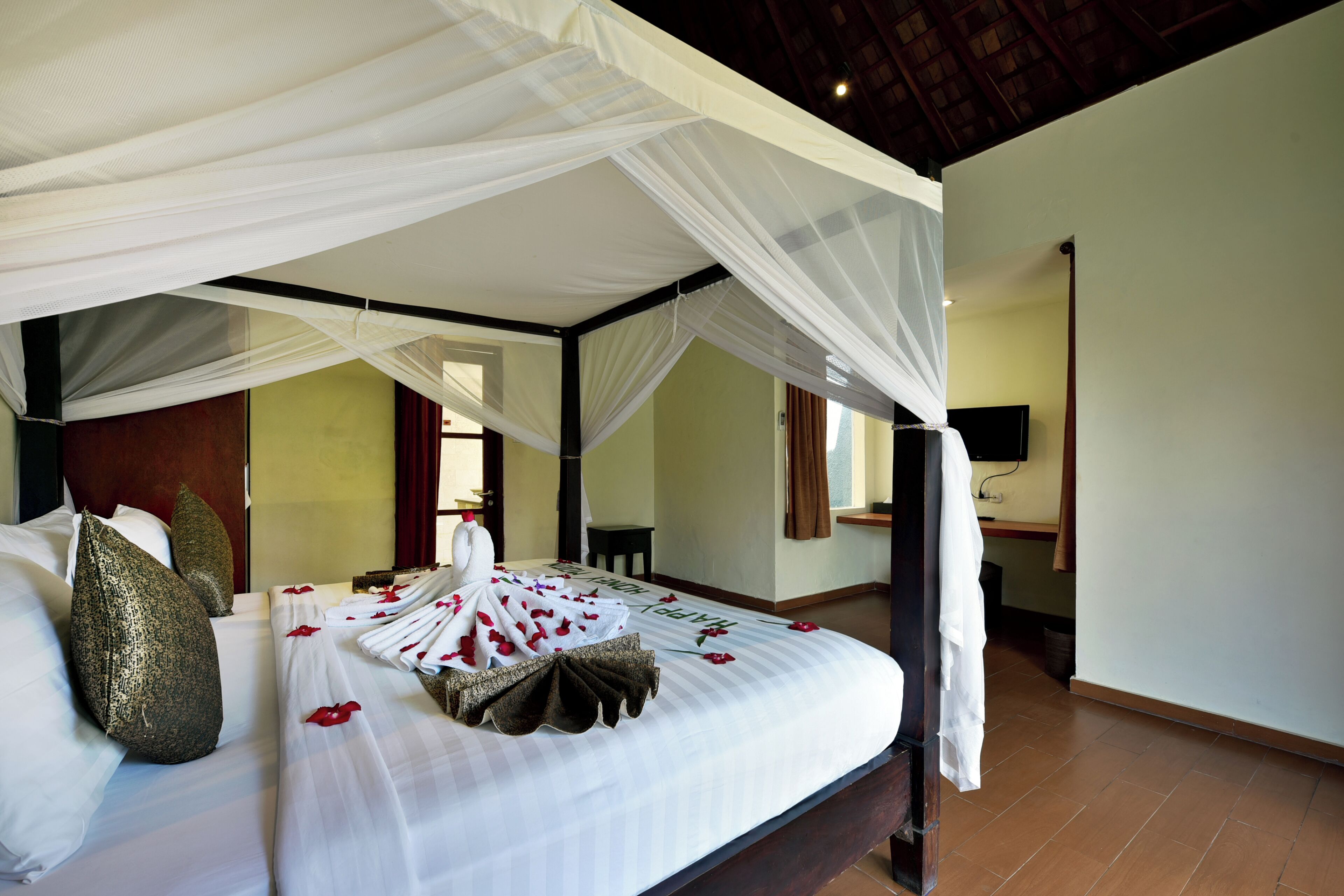 A four-poster canopy bed with white linens and rose petals, a wooden headboard, and a television on the wall.