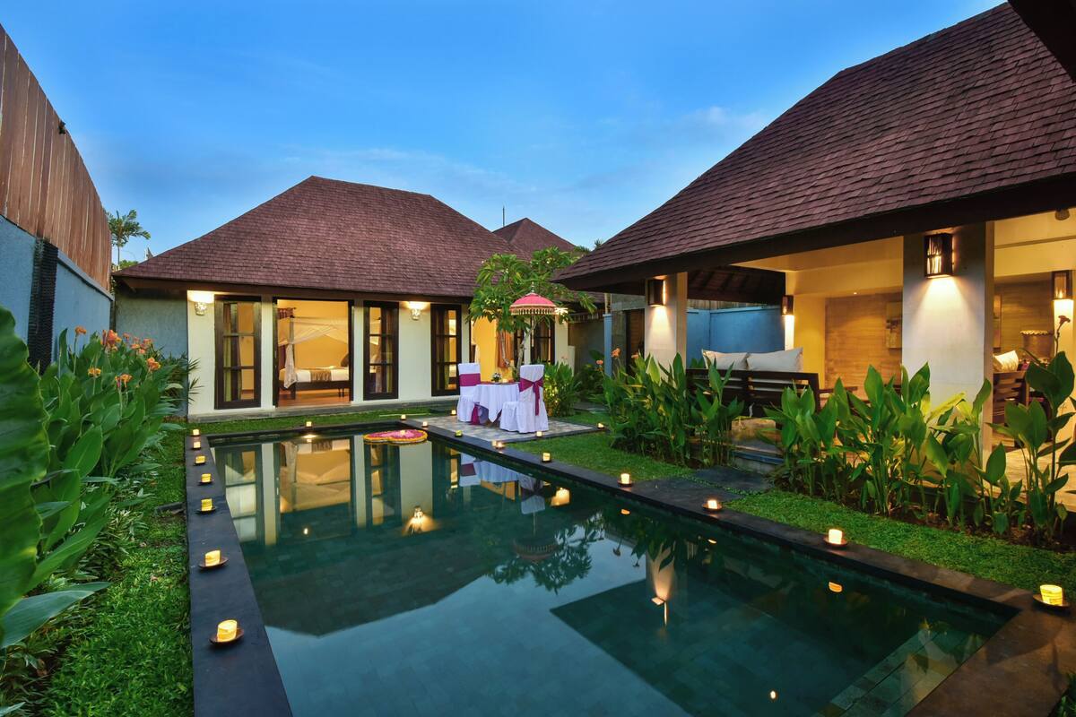 A pool area with a table set for two, surrounded by greenery and candles.