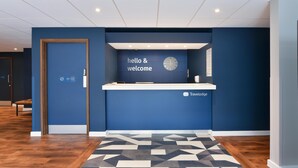 Un espace d’accueil avec un mur bleu, une horloge, une pancarte indiquant « Bonjour et bienvenue » et le logo de « Travel Lodge ».