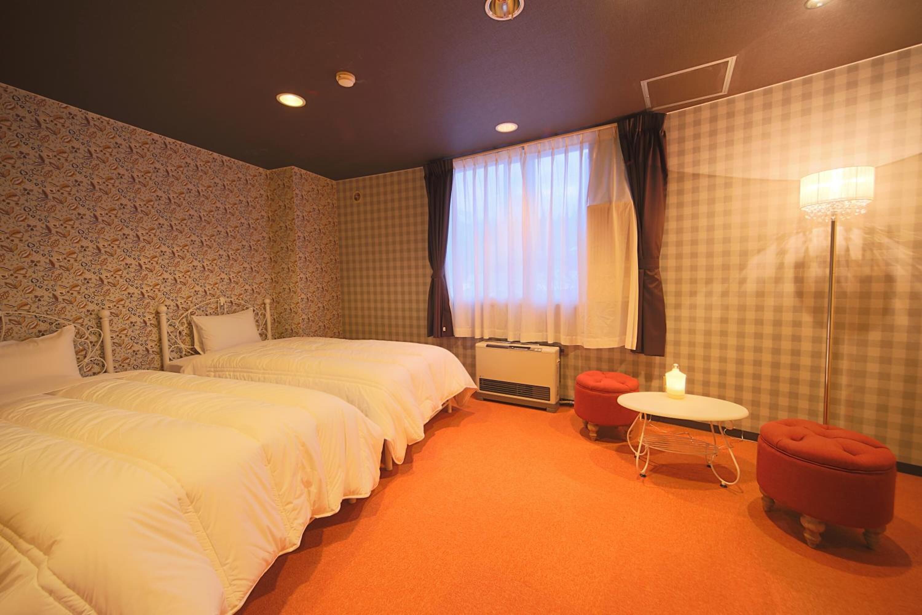 A hotel room with two beds, a small table, a red ottoman, a floor lamp, and a window with curtains.