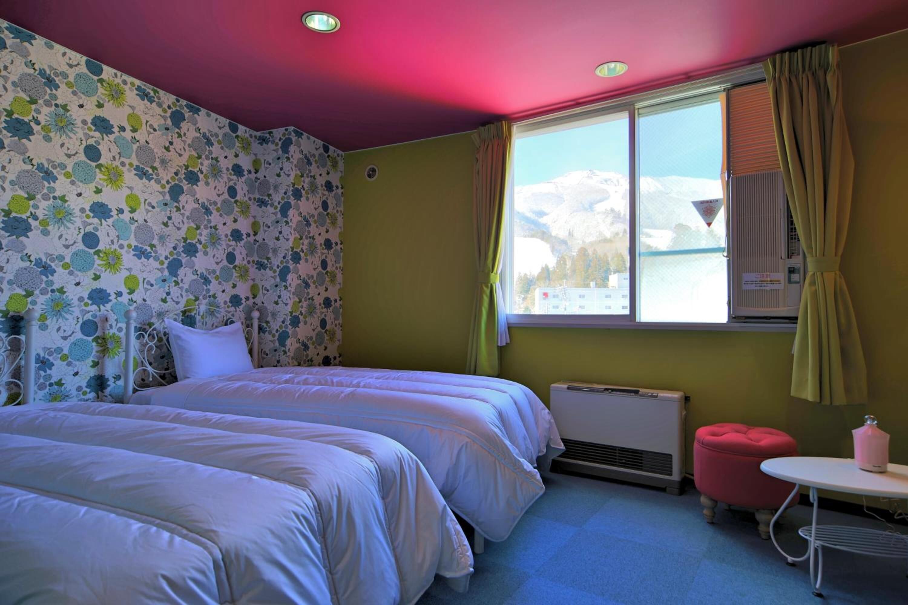 Quarto com duas camas, janela com vista para montanhas nevadas, radiador e uma pequena mesa com abajur rosa.