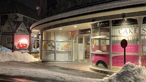 Un hotel con fachada de vidrio, señalización que dice 'Hotel 24 Sweet' y un cartel de 'CAKE S' que indica una cafetería y dulces.