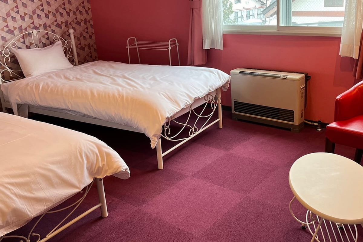 A bedroom with two beds, a red chair, a small table, and a window with curtains.