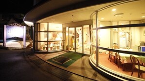 Fachada de hotel de vidrio, con área de recepción que incluye una mesa y sillas, y una escalera visible que conduce a los pisos superiores.