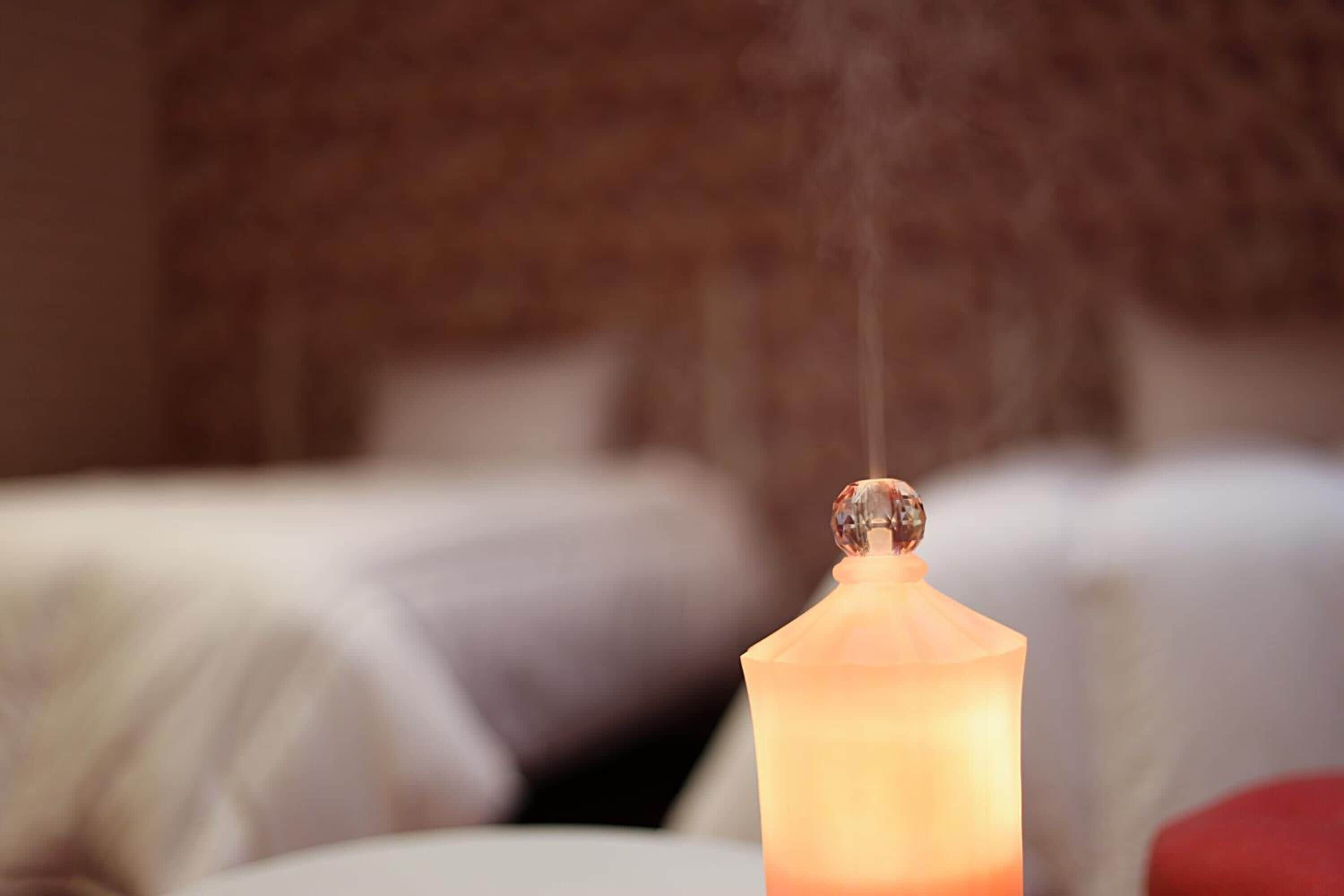A candle with smoke rising, placed on a table in a room with a bed and pillows in the background.