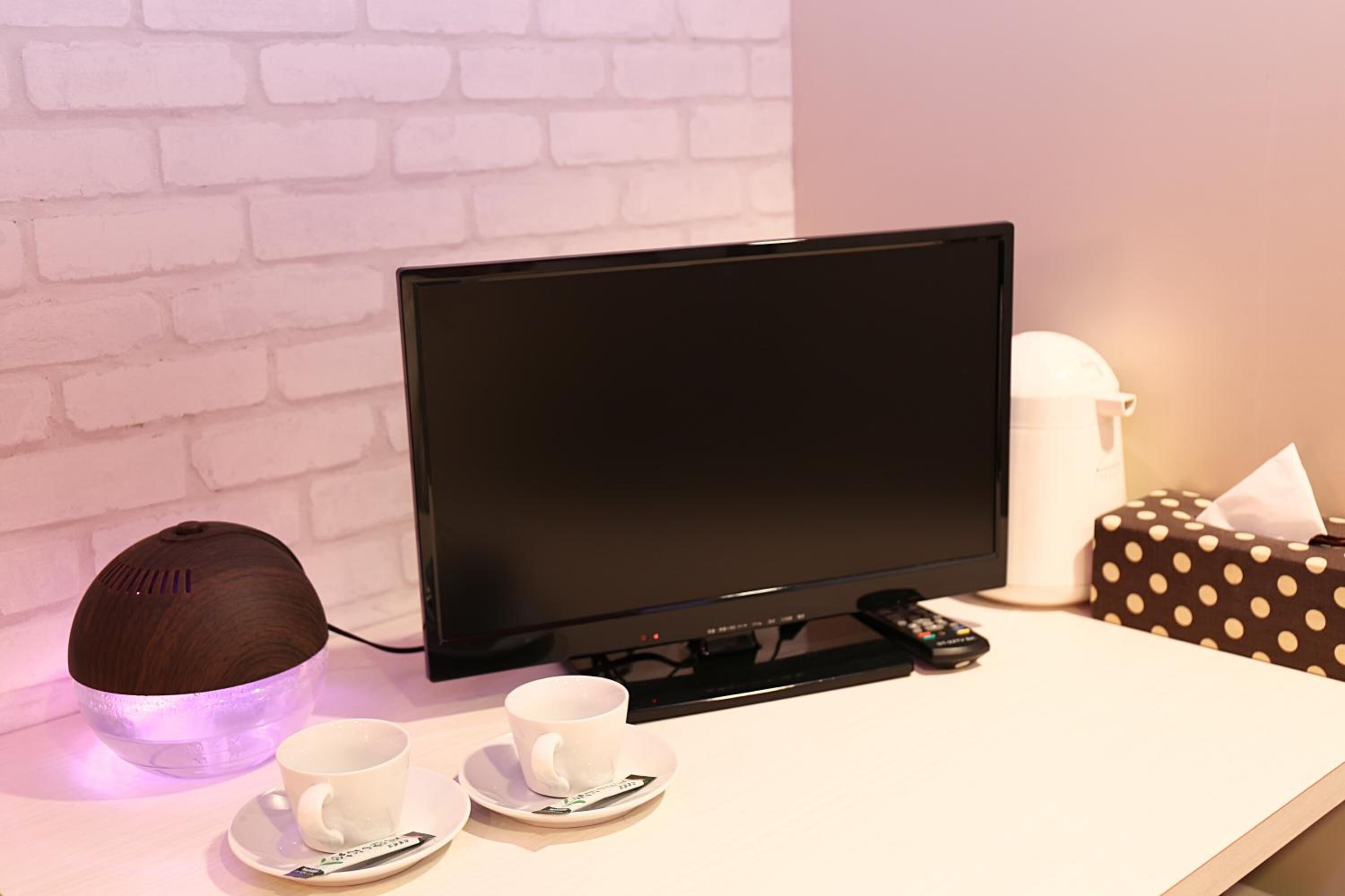 A hotel room with a flat-screen TV, a white brick wall, a white coffee table, two white cups on saucers, a brown speaker, and a polka-dotted tissue box.