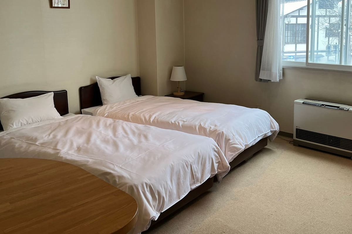 A hotel room with two beds, a round wooden table, a red ottoman, a window with curtains, and a radiator.