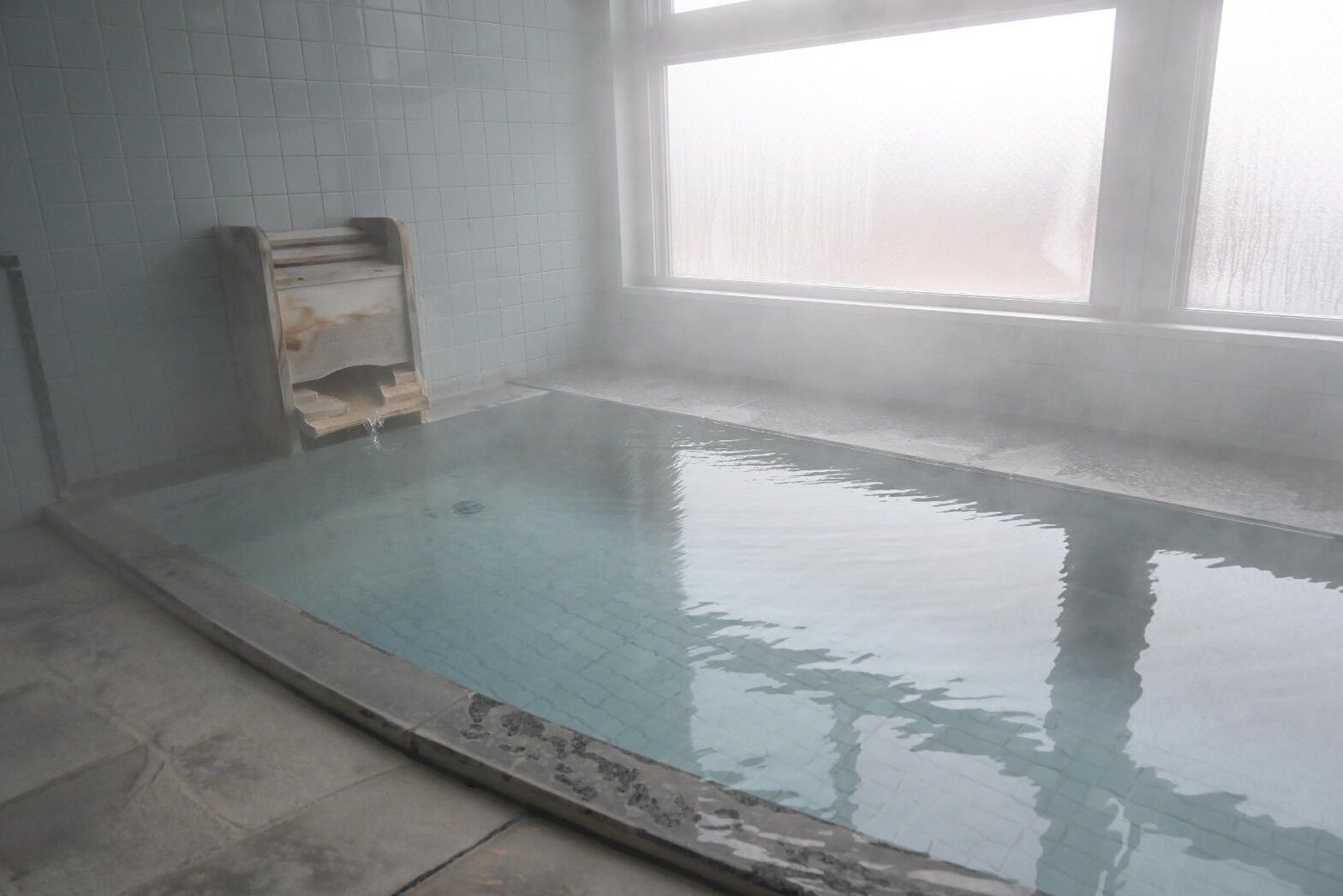 A clear indoor swimming pool with a tiled floor and walls, a window with frosted glass, and a wooden bench.