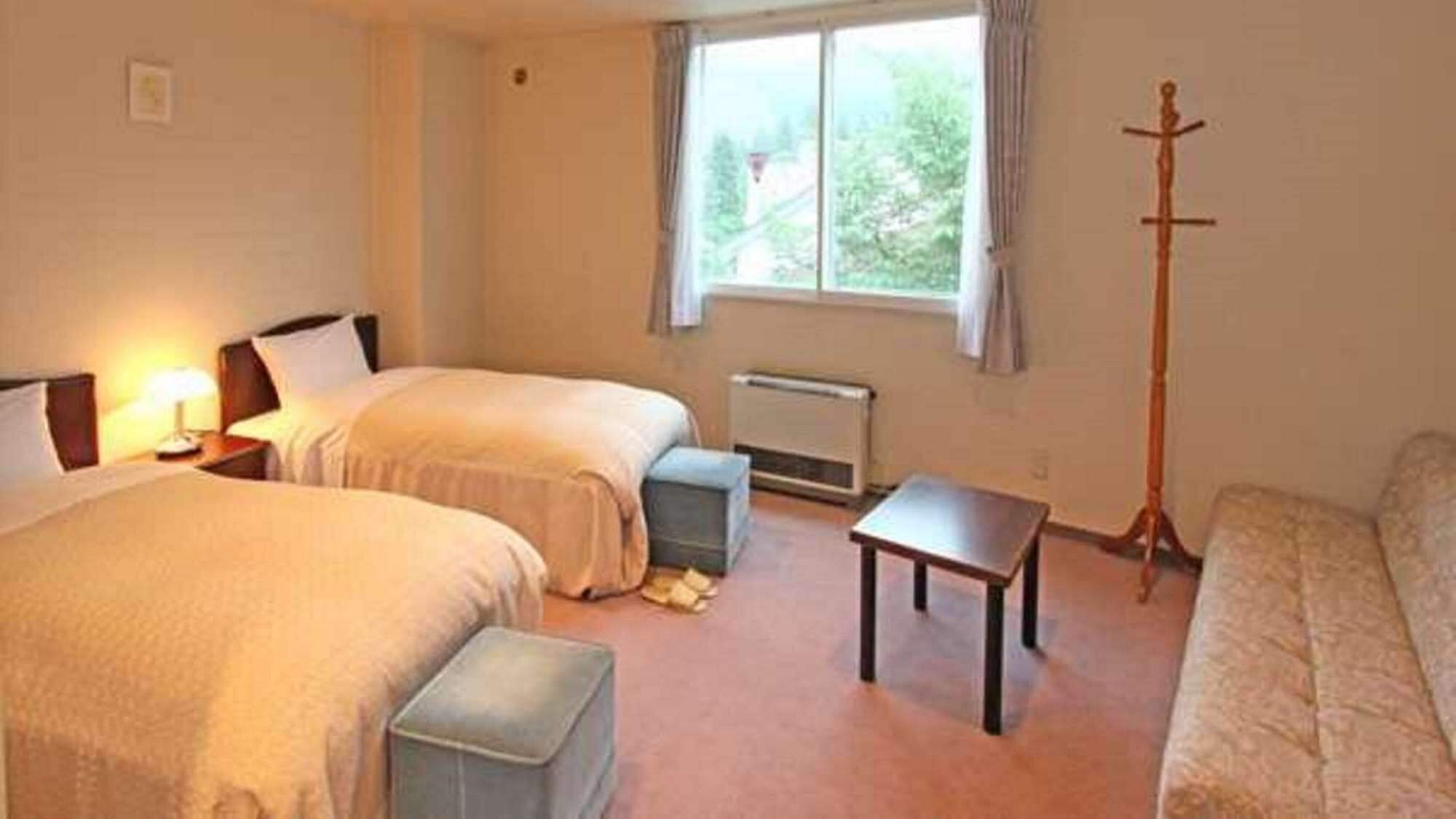 Een hotelkamer met twee bedden, een klein tafeltje, een radiator en een raam met uitzicht op bomen.