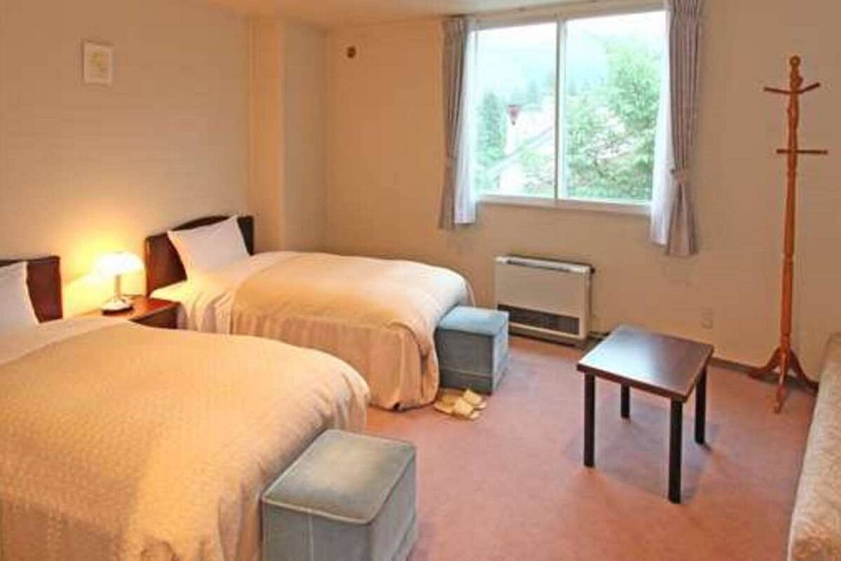 A hotel room with two beds, a small table, a radiator, and a window with a view of trees.