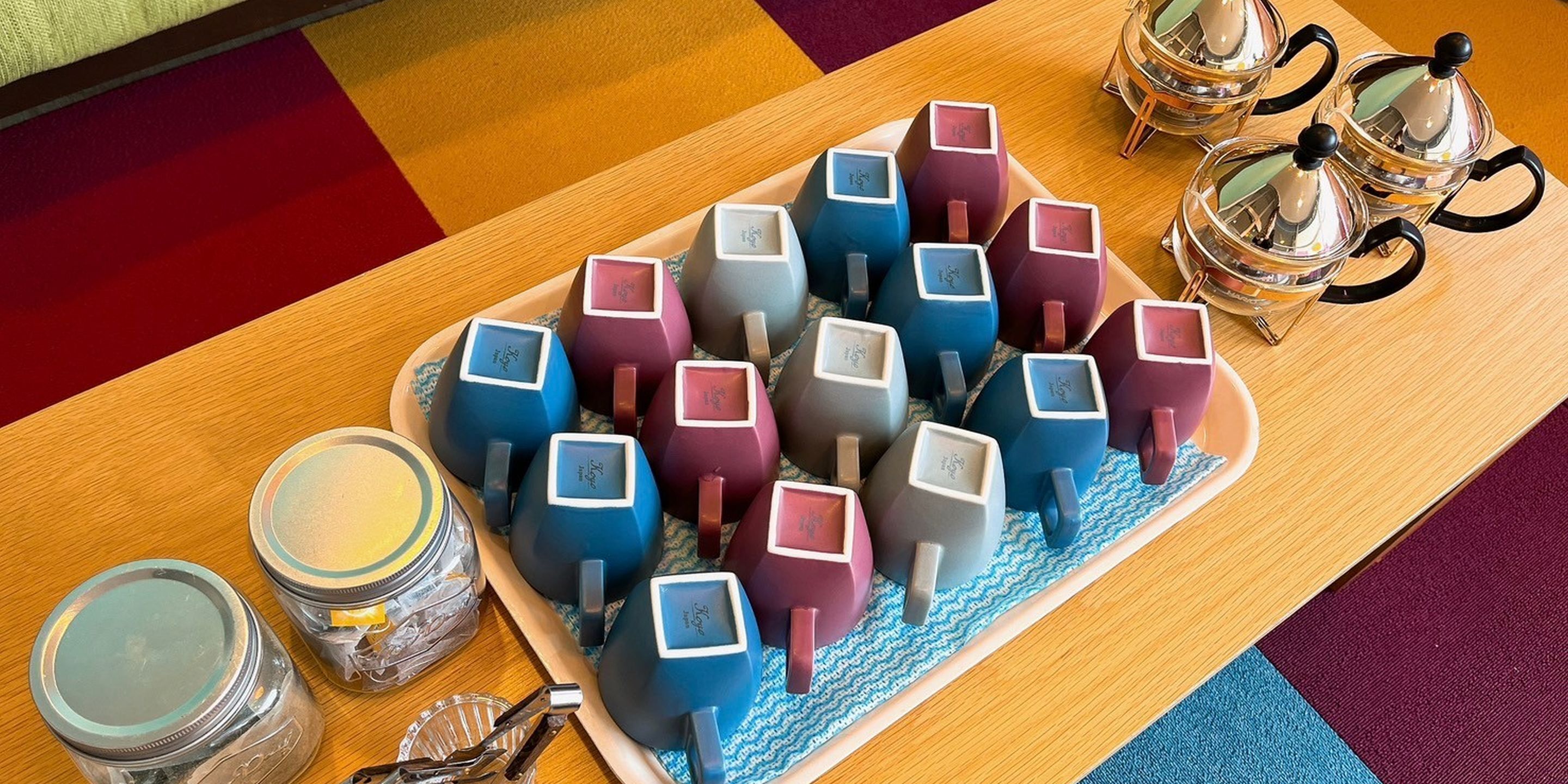 A tray with colorful mugs and two glass jars on a wooden table.