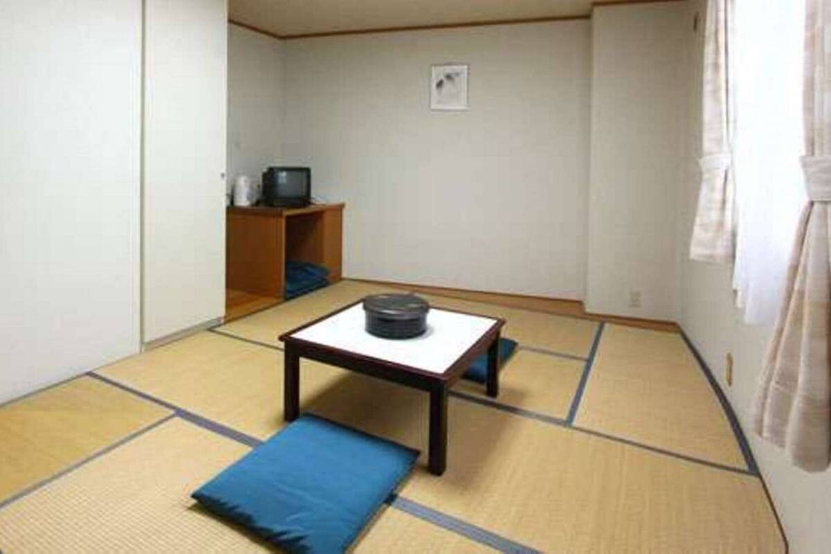 A traditional Japanese room with tatami flooring, a low table with a cushion, and a television on a shelf.