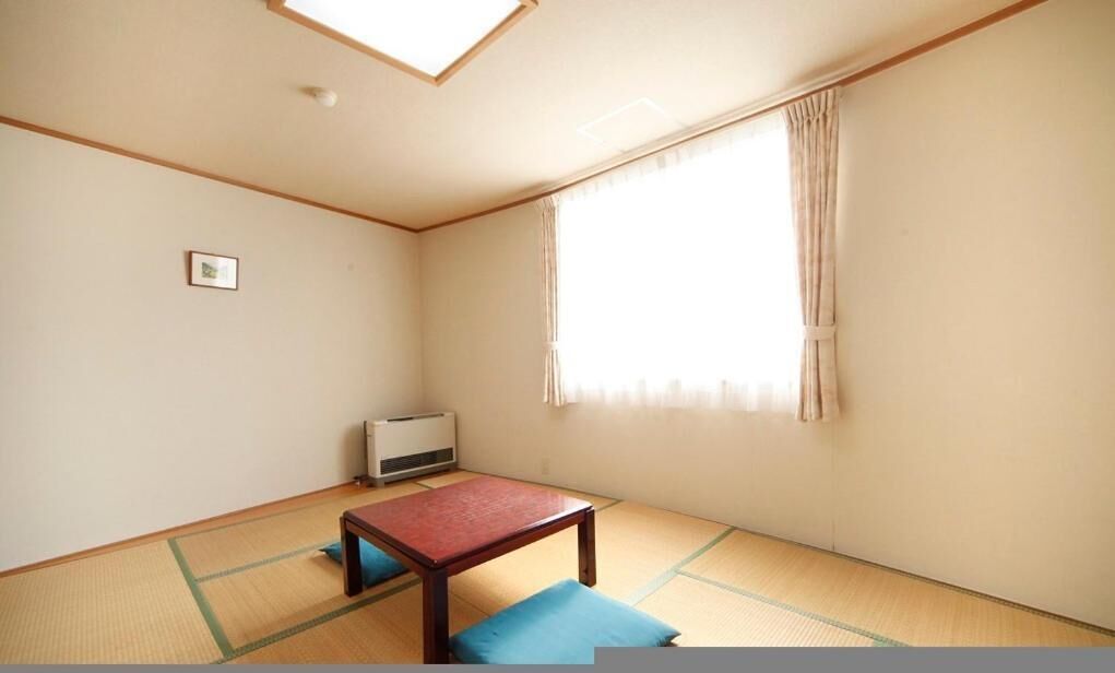 Una habitación con suelo de tatami, una mesa de madera baja, un cojín azul, un calentador y una ventana con cortinas.