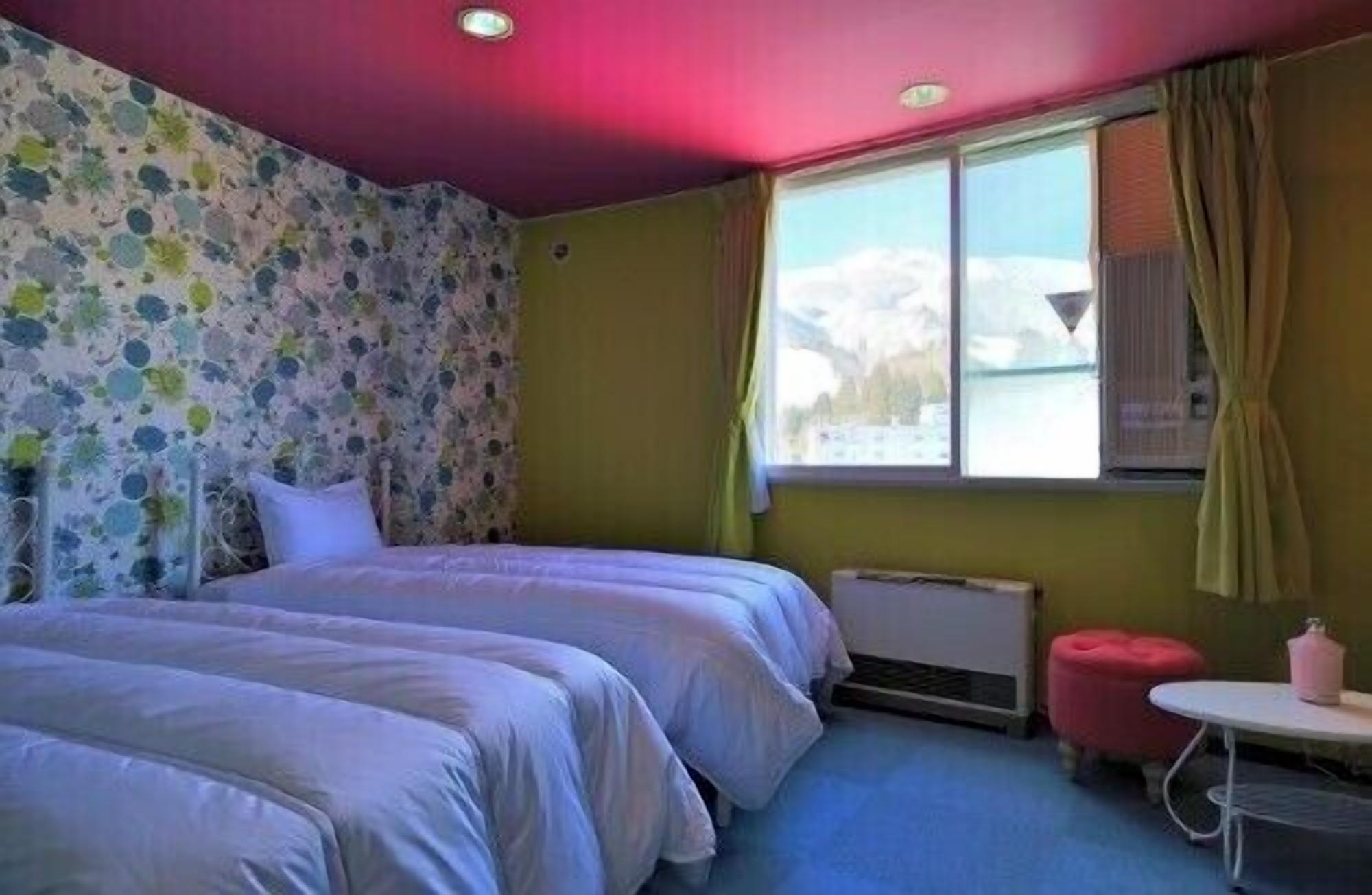 Een kamer met twee bedden, een raam met gordijnen, een radiator en een klein tafeltje met een roze stoel.