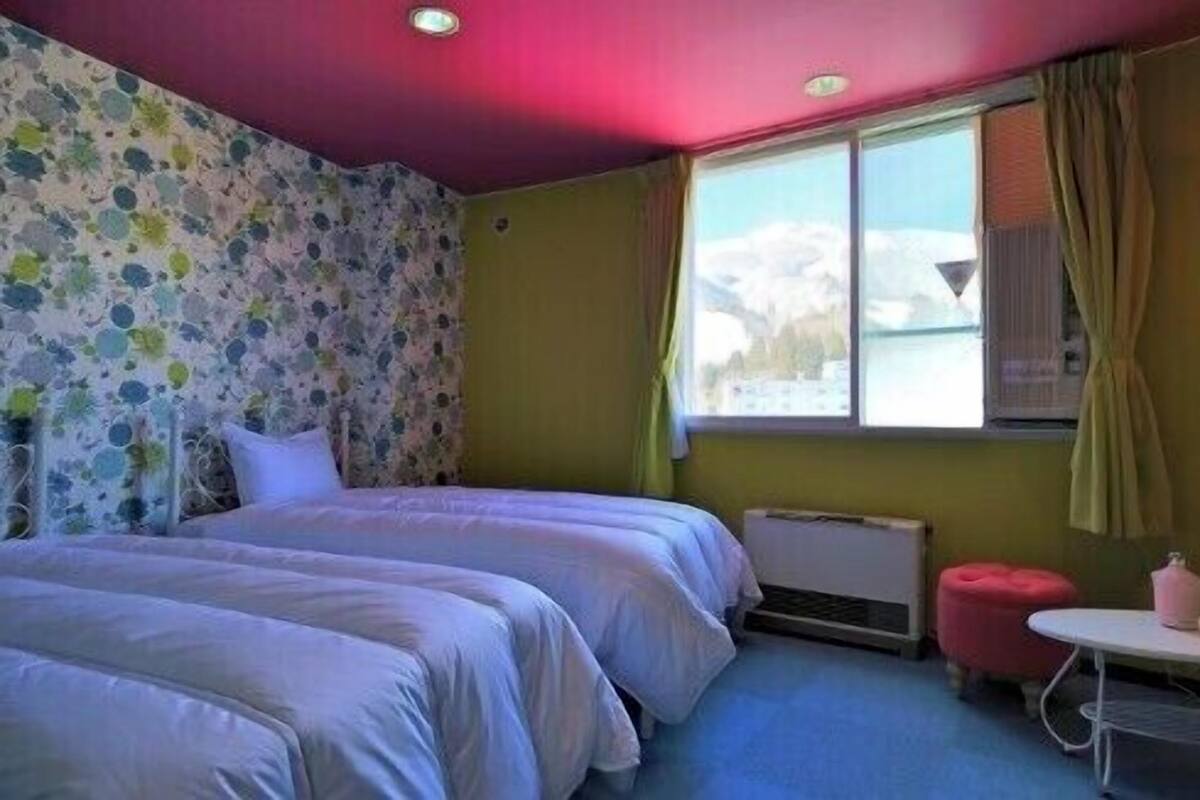 A room with two beds, a window with curtains, a radiator, and a small table with a pink chair.