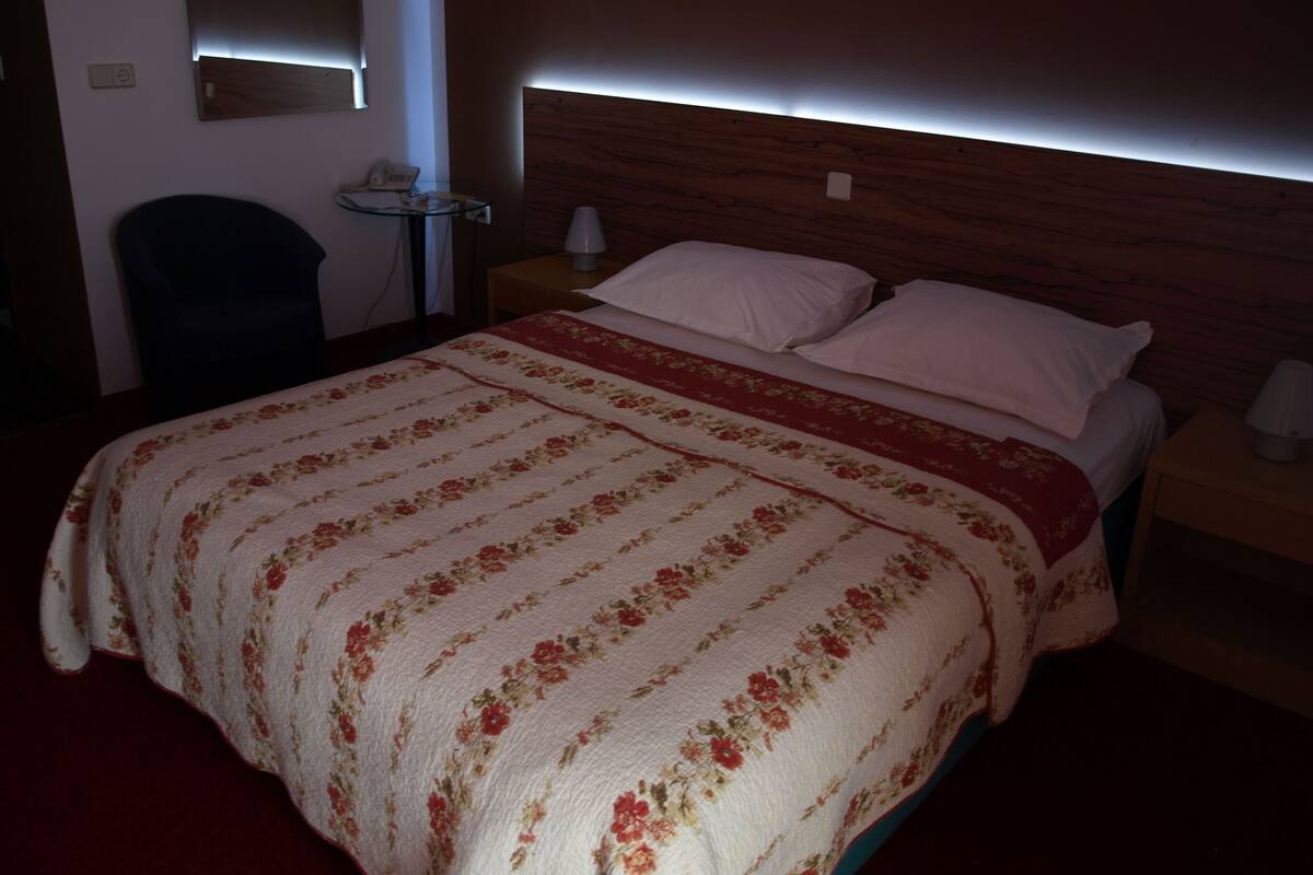Une chambre d’hôtel avec une tête de lit en bois, une couvre-lit à motifs floraux, deux oreillers blancs, une table de chevet avec une lampe, et une petite table ronde avec un téléphone et une lampe.
