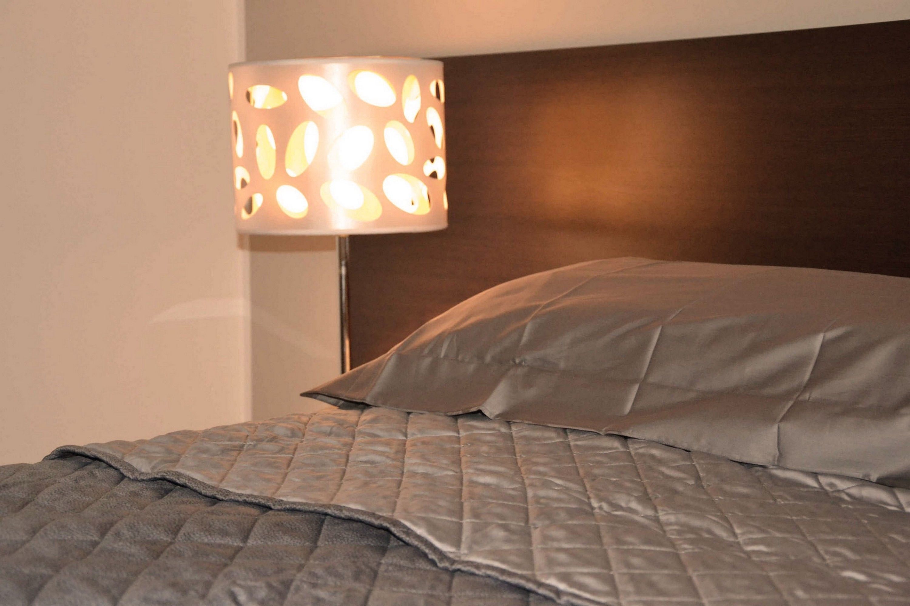 Una cama con un edredón acolchado y una almohada, una lámpara de pared con pantalla estampada y un cabecero de madera.