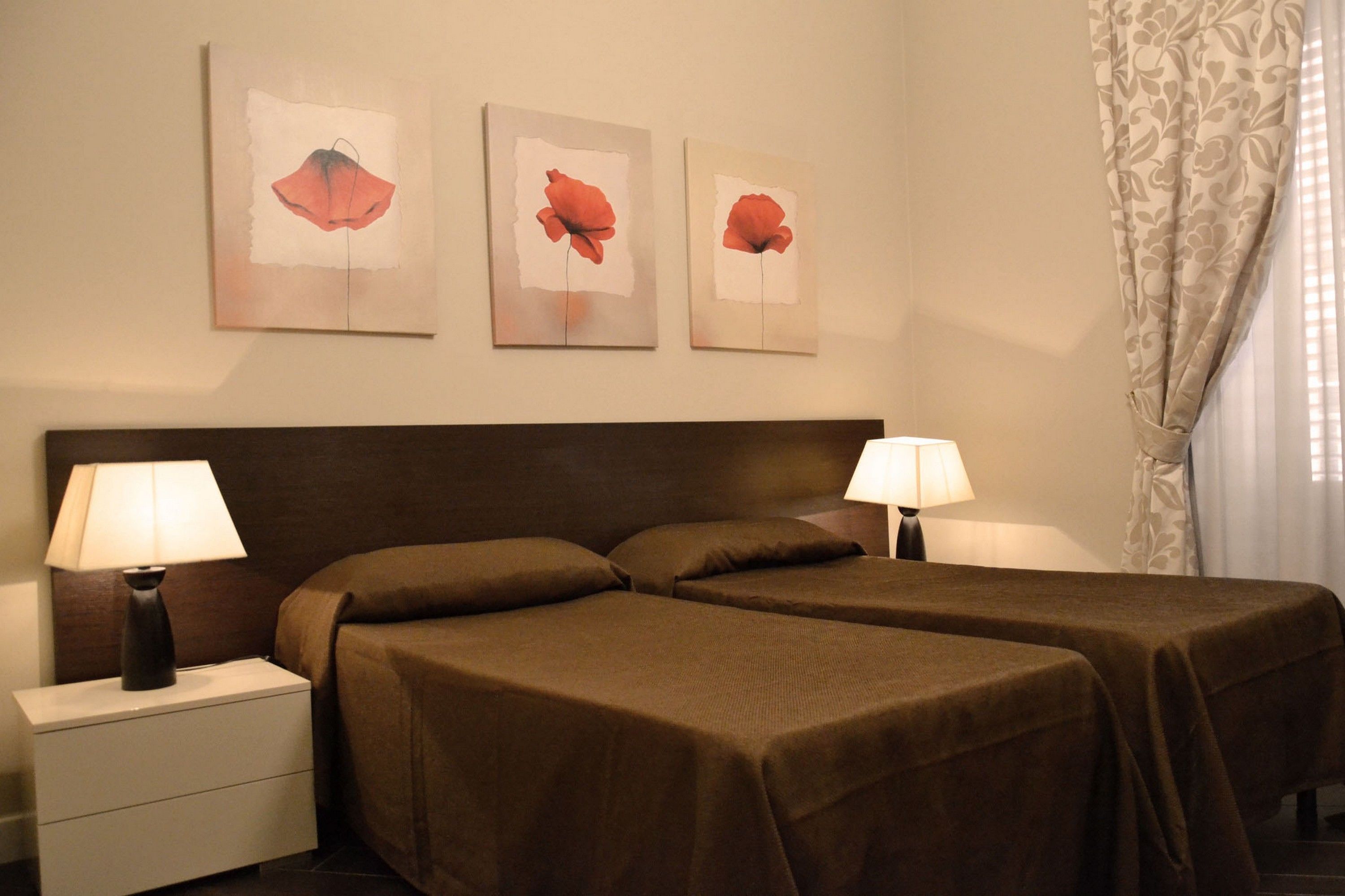 Une chambre d’hôtel avec deux lits, une table de chevet avec des lampes et trois tableaux représentant des fleurs rouges, accrochés au mur.