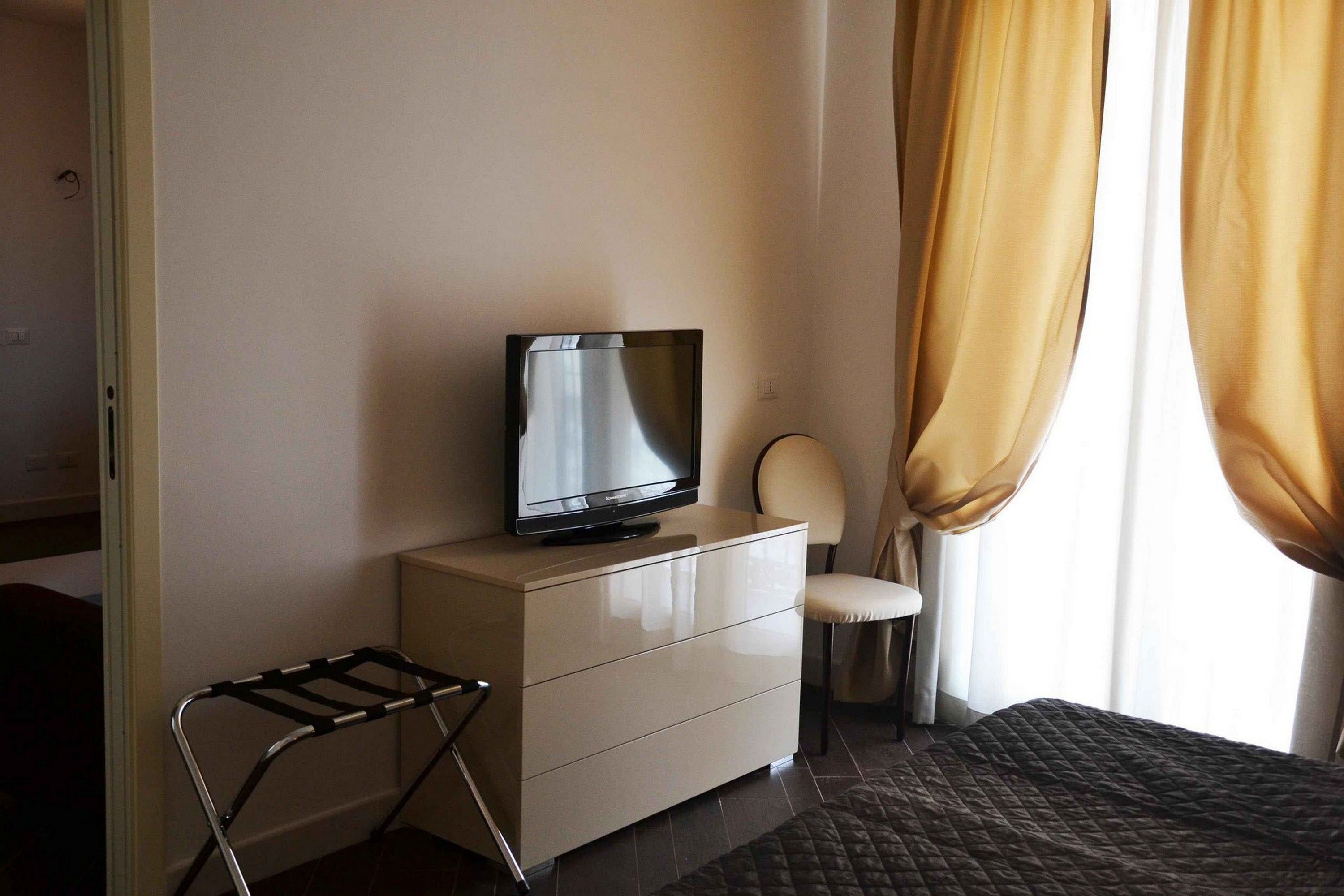 Ein Hotelzimmer mit Fernseher auf einem Schminktisch, einem Sessel und einem Fenster mit Vorhängen.