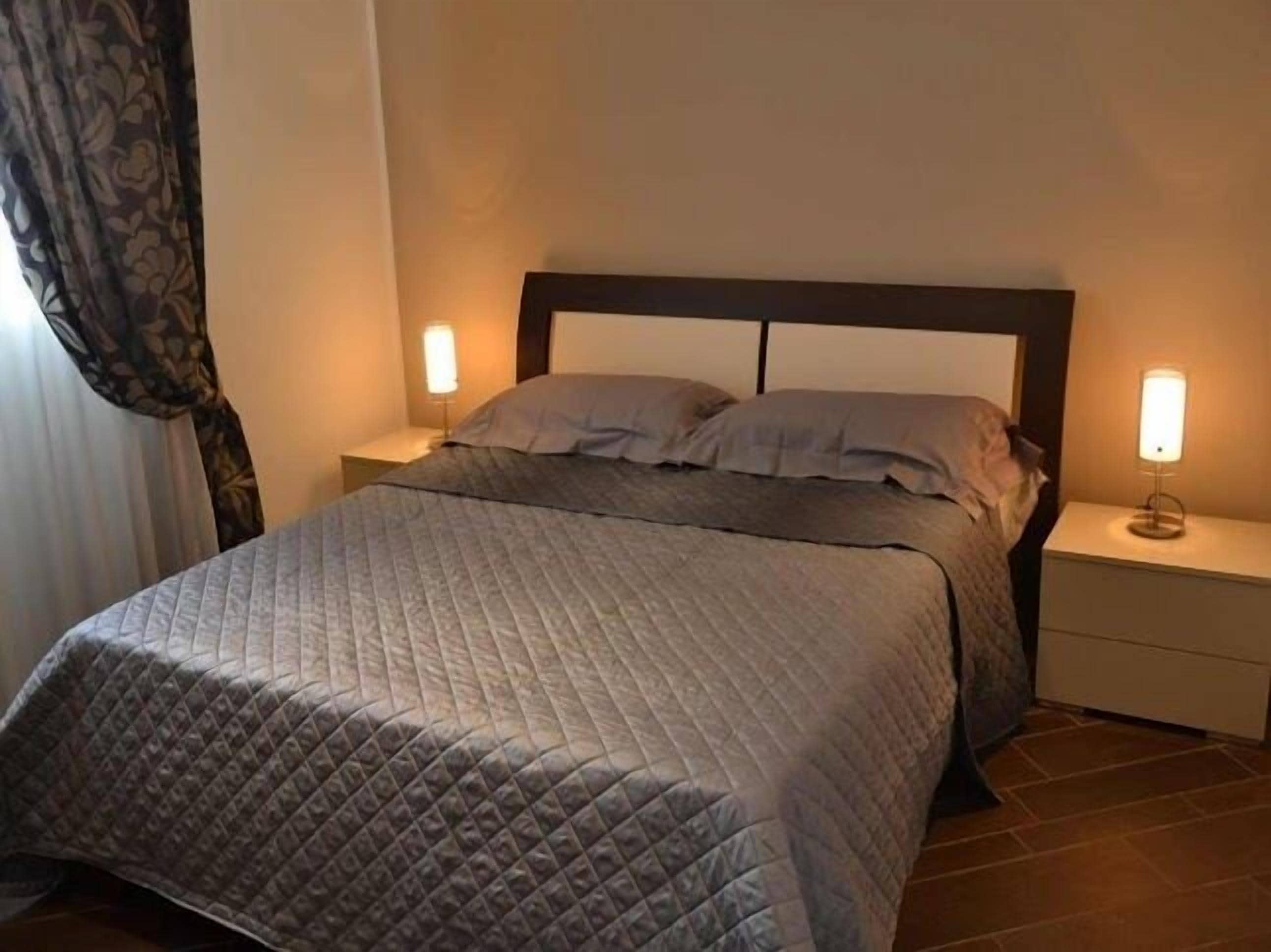 A neatly made bed with a quilted cover, two bedside tables with lamps, and a window with curtains.