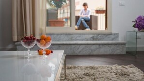 A room with a glass table displaying fruit bowls, a view of a balcony with a person sitting, and a potted plant.