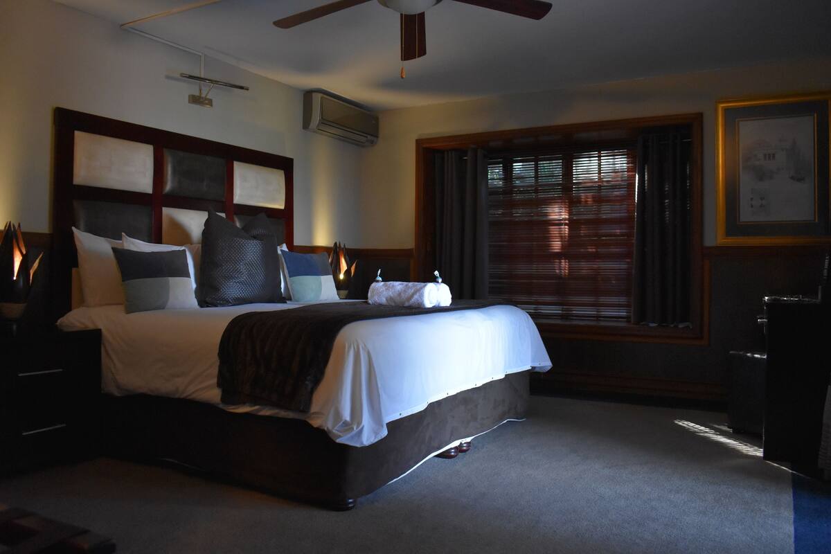 A well-lit hotel room with a large bed, a ceiling fan, a window with blinds, and a framed picture on the wall.