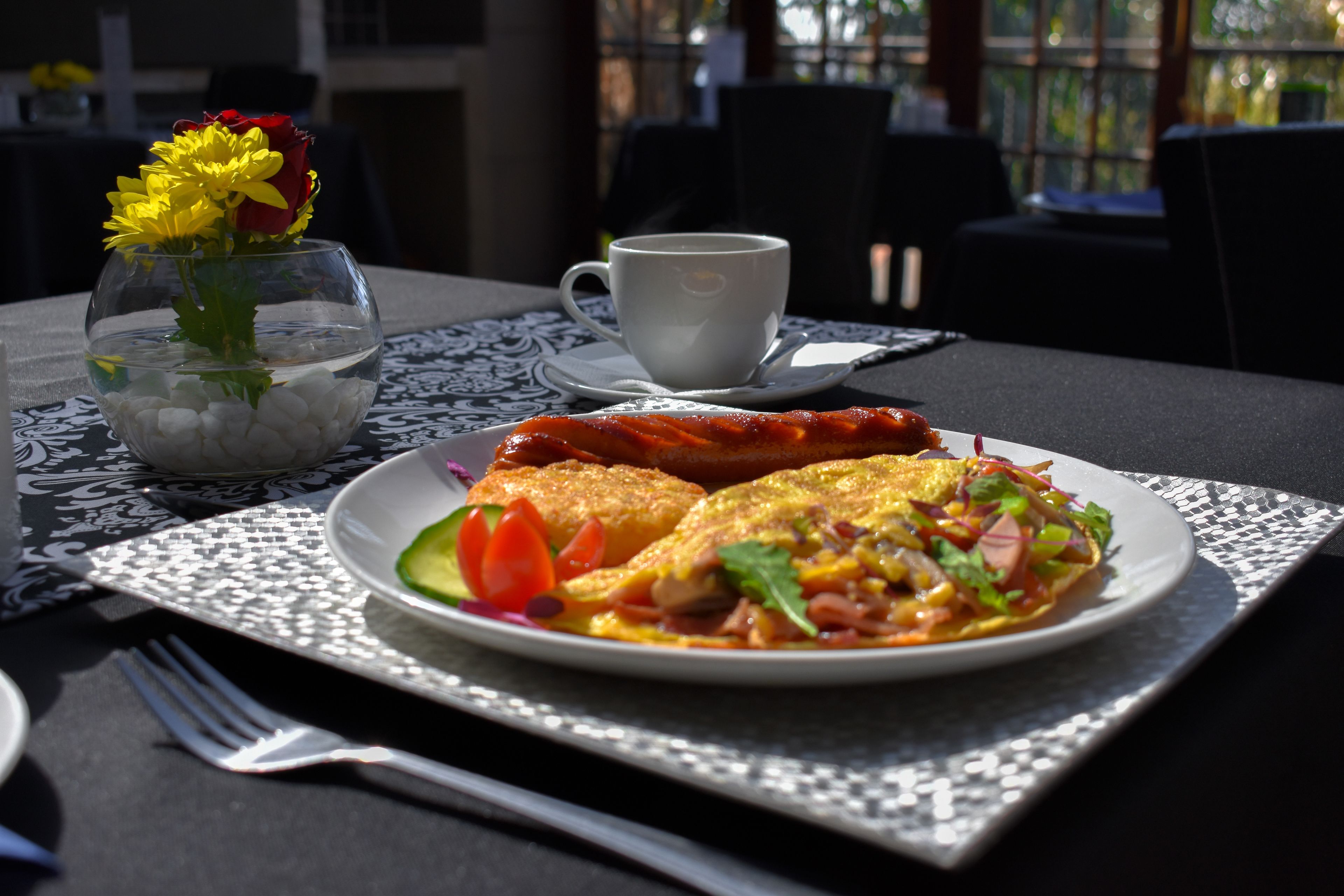 Une table à manger avec une assiette de nourriture, une tasse de café et un vase rempli de fleurs.