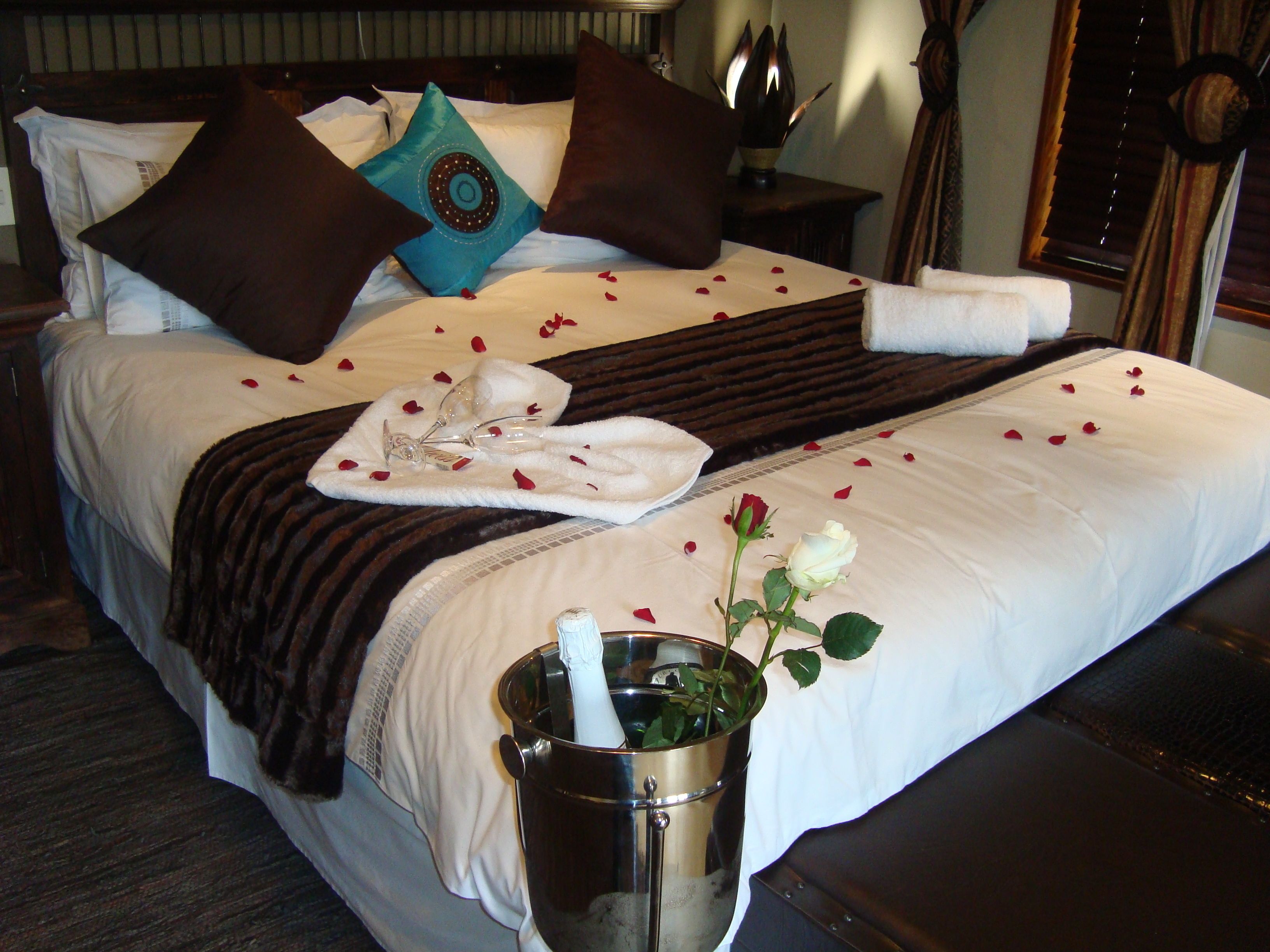 Ein ordentlich gemachtes Bett mit weißer Decke, braunen Kissen und einem blauen Zierkissen. Rosenblätter sind auf dem Bett verteilt und ein weißes Handtuch liegt darüber gefaltet. Eine weiße Rose steht in einem Metallkübel.