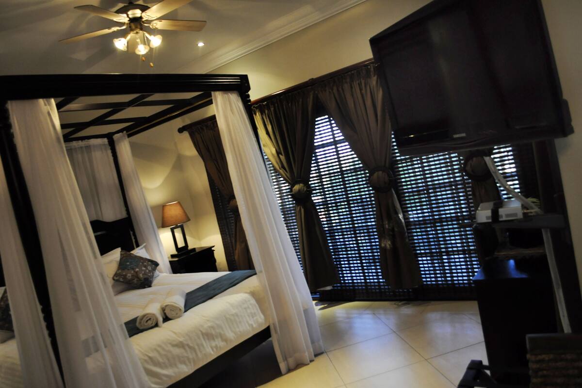 A four-poster bed with white linens, a ceiling fan, and a TV on a stand.