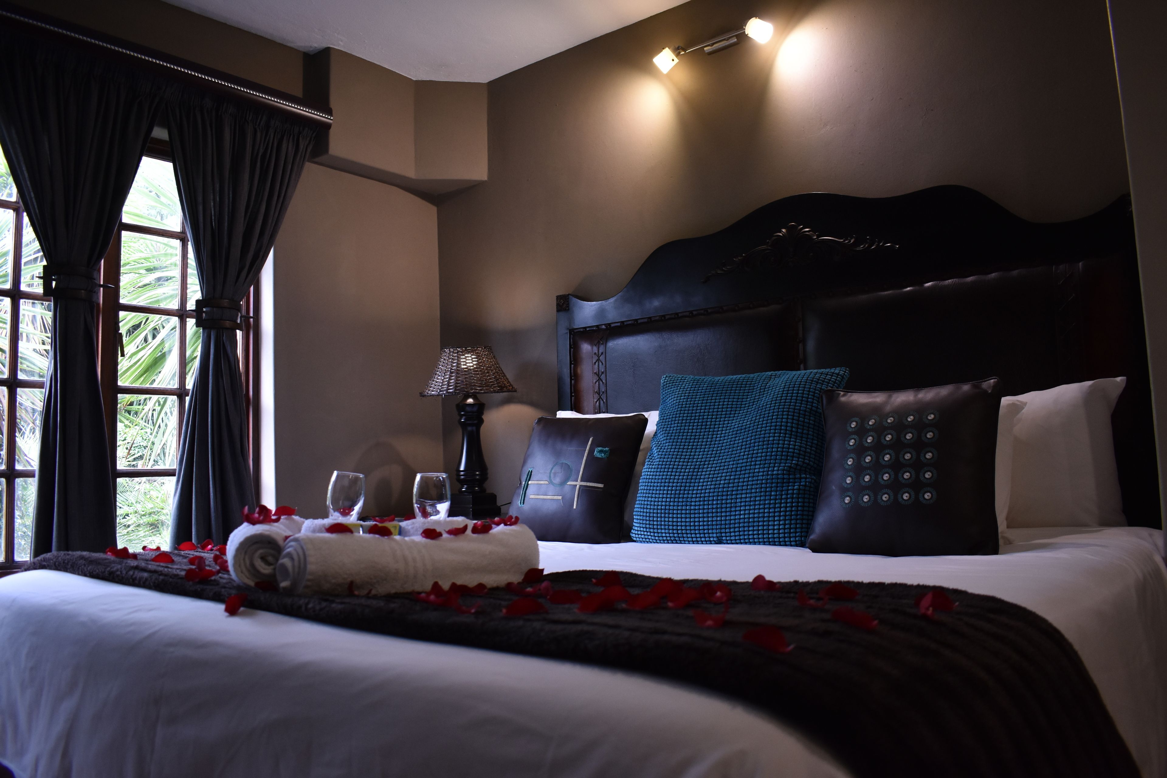 A hotel room with a large bed, a nightstand, a lamp, and a view of palm trees through the window.