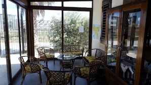 Un porche cubierto con mobiliario de mimbre y una mesa de vidrio, con vista a un jardín y una calle.