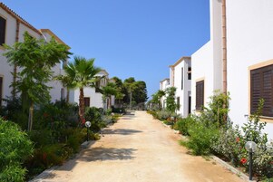 Una stretta via alberata con edifici bianchi e persiane scure, che evoca un’ambientazione mediterranea.