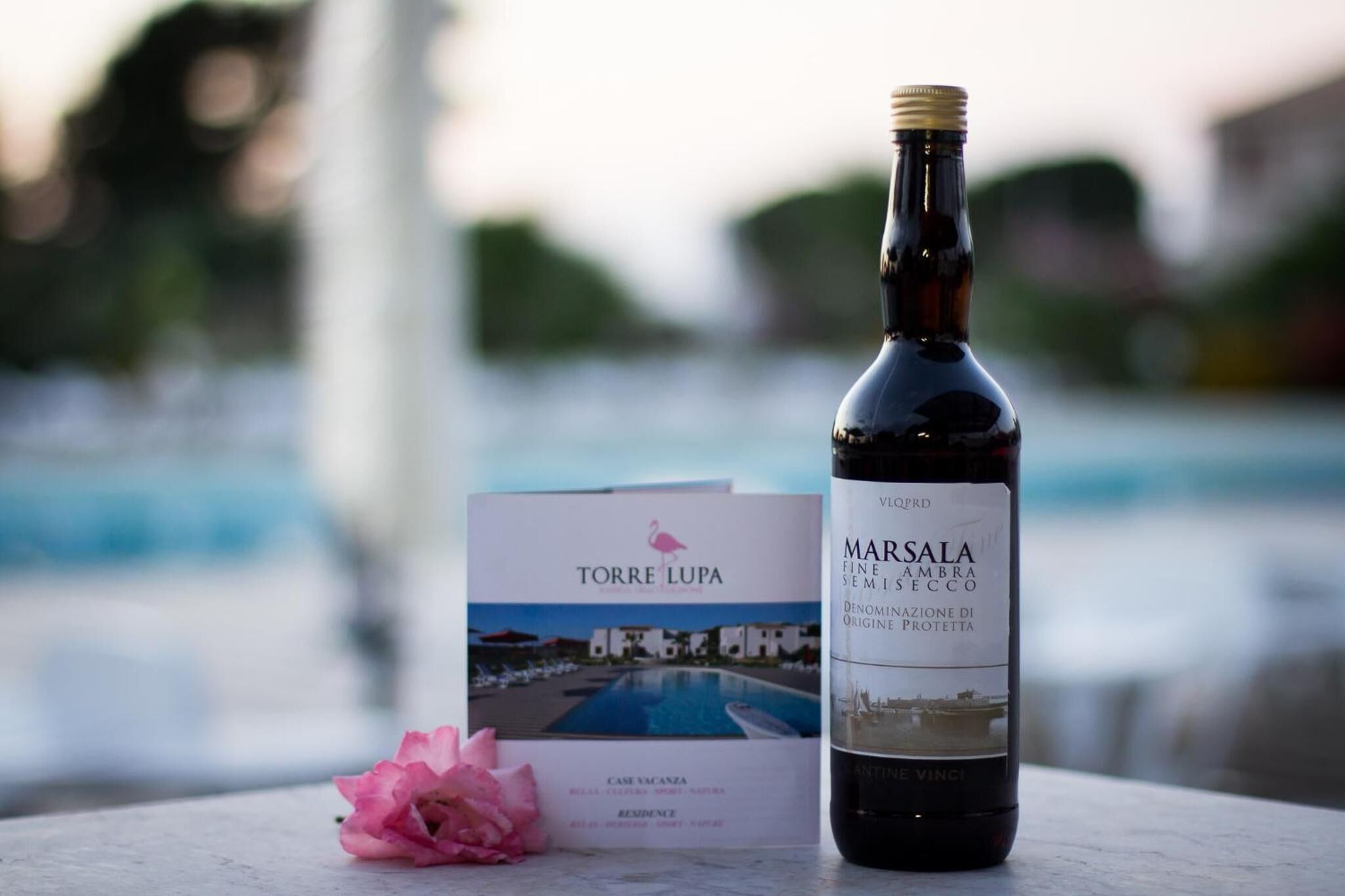 Una bottiglia di vino Marsala, un depliant per Torre Lupa e un fiore rosa su un tavolo vicino a una piscina.