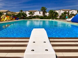 Una piscina con una poltrona da sdraio bianca in primo piano, palme e un edificio con tetto in tegole sullo sfondo.