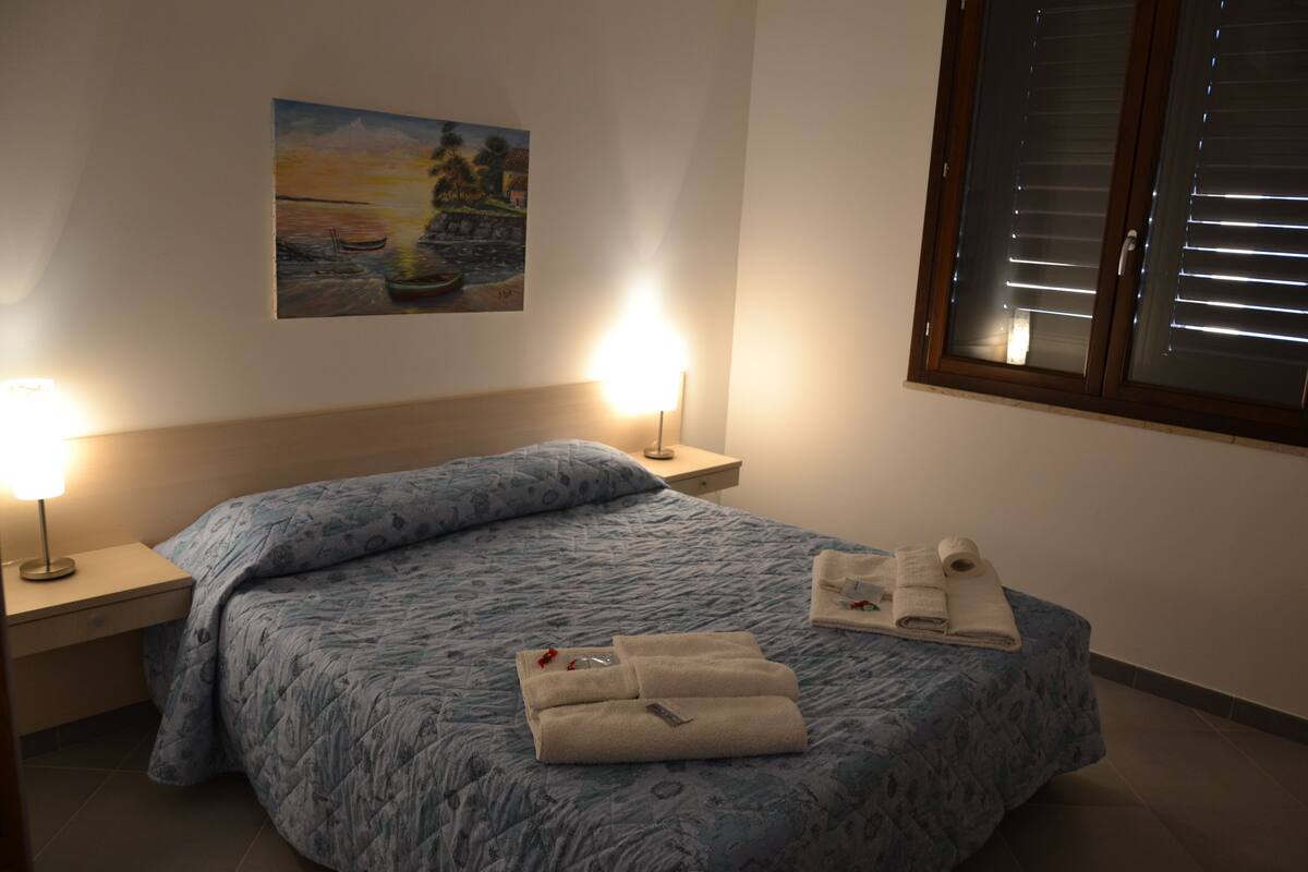 A neatly made bed with a patterned blue bedspread, two bedside lamps, a painting on the wall, and a window with blinds.