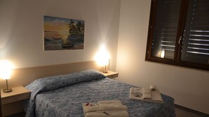 Una cama bien hecha con una colcha azul estampada, dos lámparas de noche, un cuadro en la pared y una ventana con persianas.