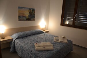 Cama bem arrumada com colcha azul estampada, duas luminárias de cabeceira, um quadro na parede e uma janela com persianas.