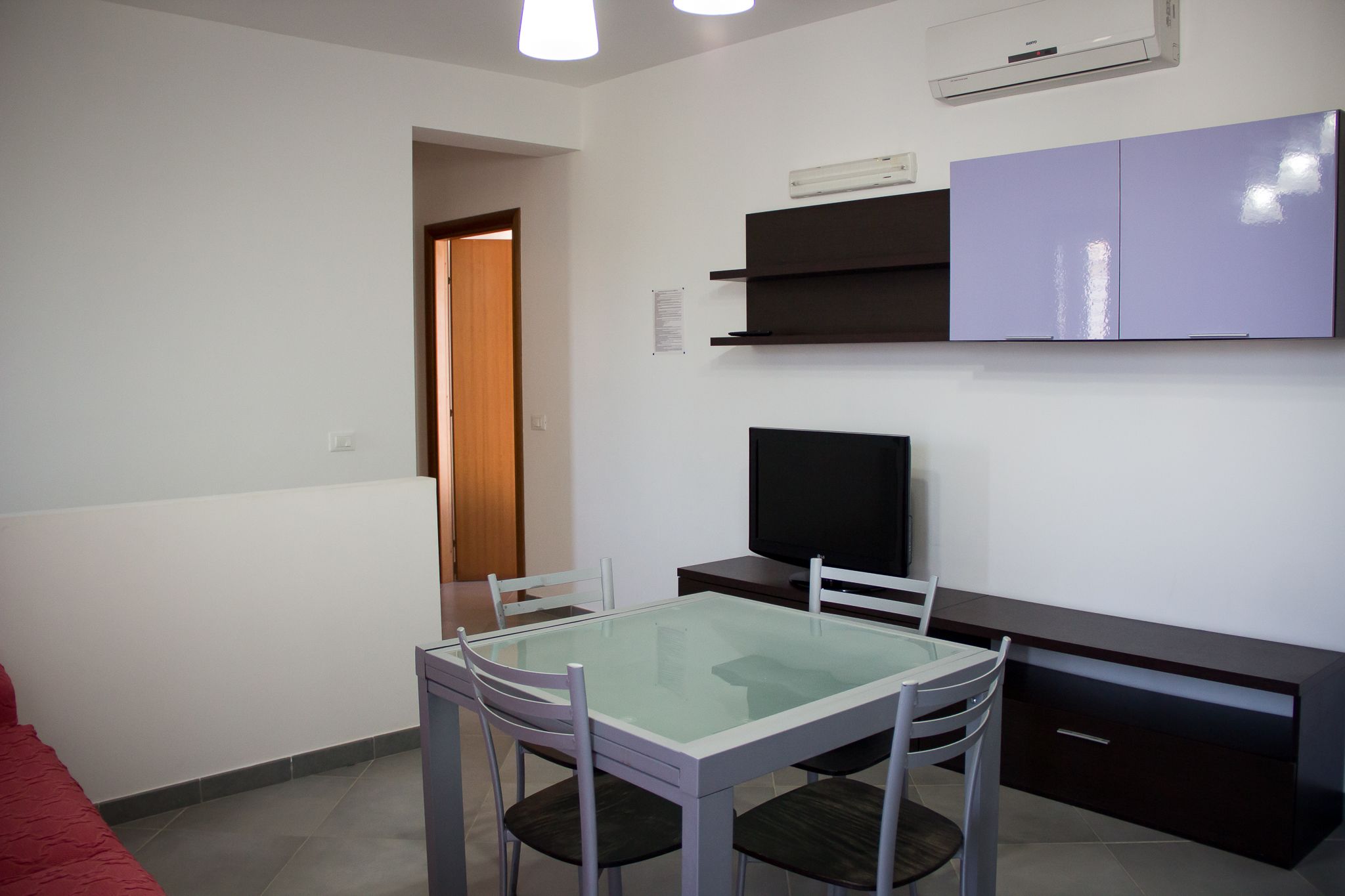 Una habitación pequeña y moderna con una mesa de comedor de cristal, un televisor de pantalla plana, un escritorio de madera oscura y un armario morado.