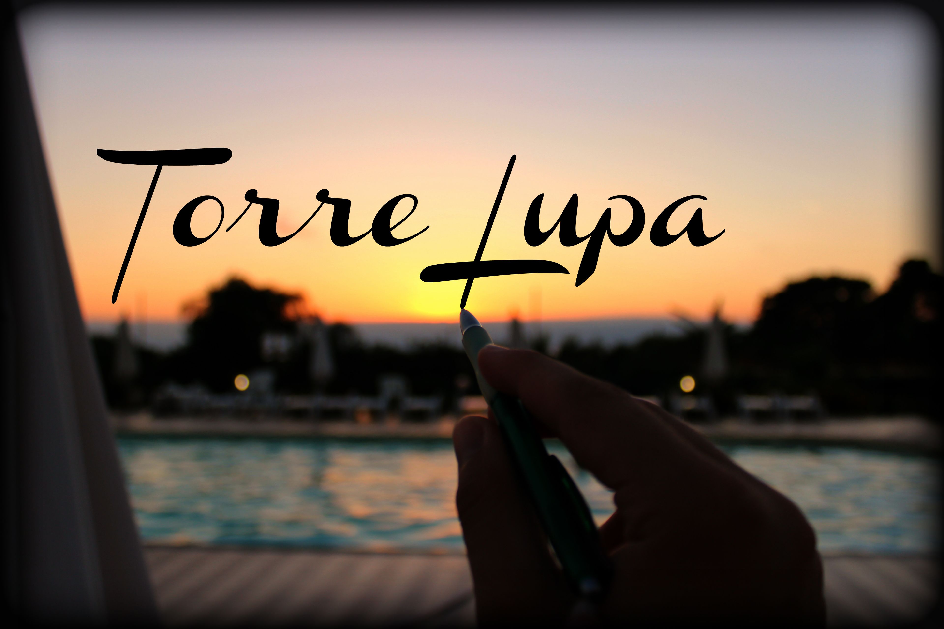 En hånd holder en pen med teksten "Torre Lupa" foran en solnedgang.