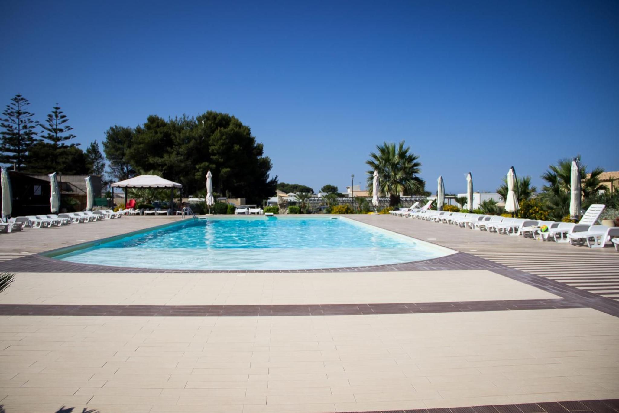 A rectangular swimming pool with a tiled deck surrounding it, lounge chairs, and umbrellas.