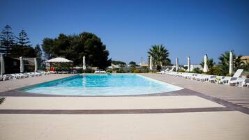 A rectangular swimming pool with a tiled deck surrounding it, lounge chairs, and umbrellas.
