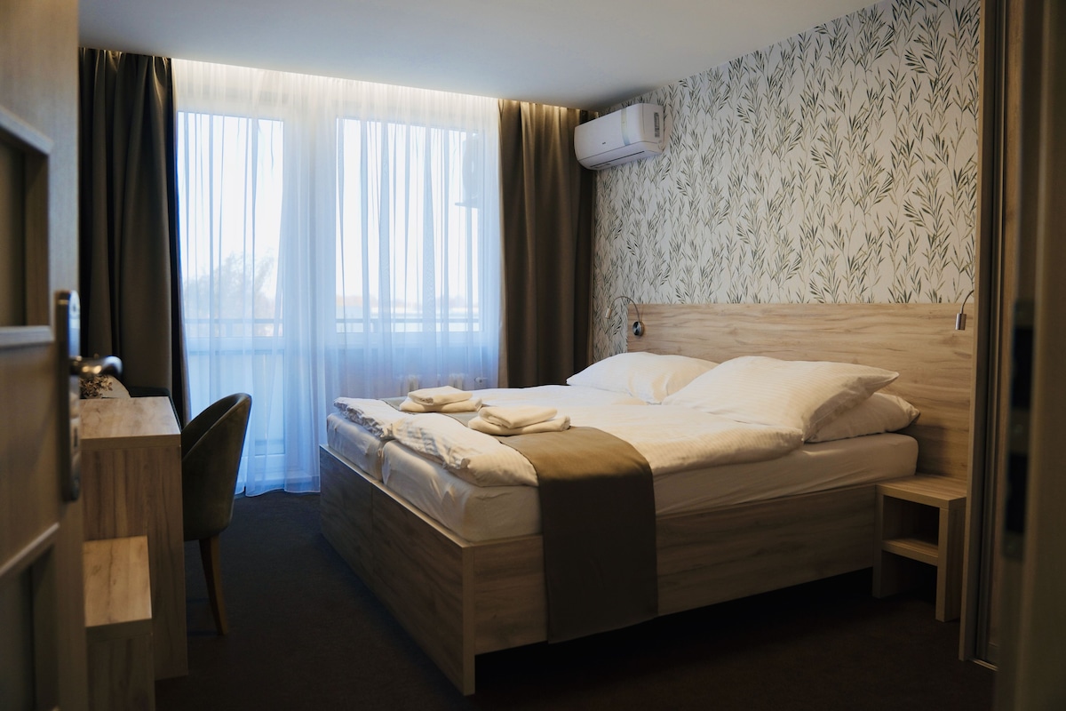 A hotel room with a bed, a desk, a chair, a window with curtains, and an air conditioning unit on the wall.