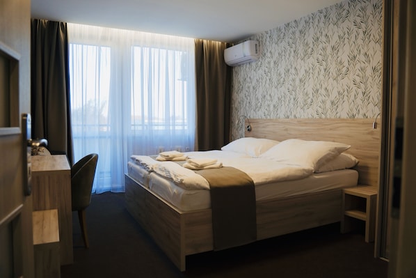 A hotel room with a bed, a desk, a chair, a window with curtains, and an air conditioning unit on the wall.