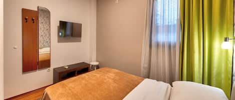 A hotel room with a bed, a TV, an air conditioner, a mirror, and a window with curtains.