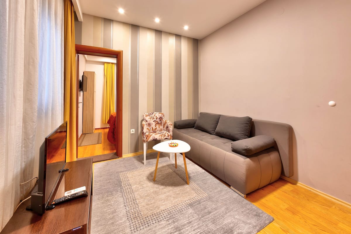 A modern living room with a grey sofa, a patterned armchair, a small white table, and a remote control on a wooden stand.