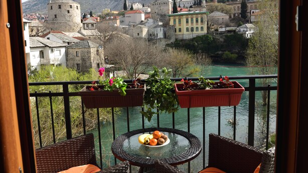 A balcony with wicker furniture, a glass table with fruit, and a view of a town with historic buildings and a river.